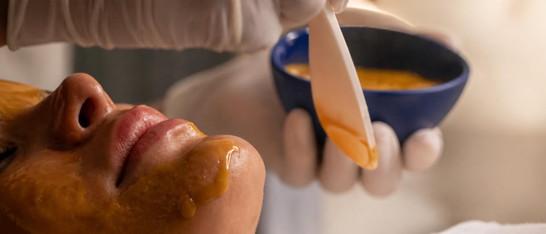 Close-up view of a person receiving a creamy DIY honey face mask, partially visible face while a gloved hand applies the mask with a spatula froma bowl.